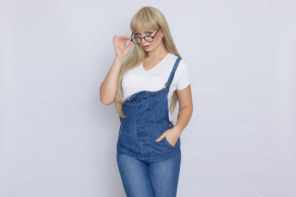 Seriously blond woman in jeans overalls holding glasses on her face over grey background - Foto, afbeelding