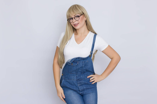 Beautiful blond longhaired woman in jeans overalls and glasses over grey background - Foto, immagini