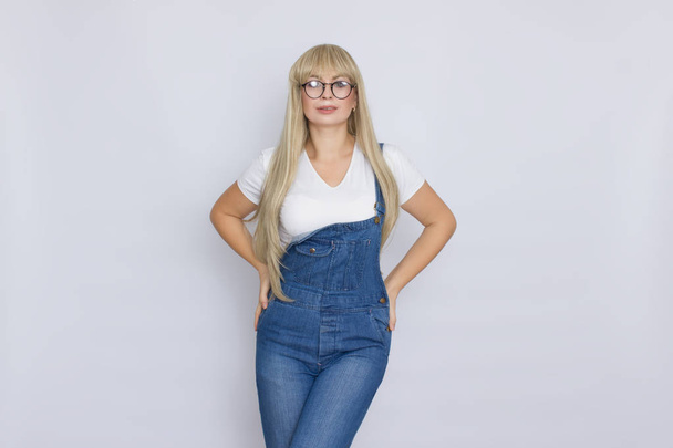 Sweet beautiful blond woman with very long hair in blue jeans overalls and glasses over grey background - Foto, afbeelding