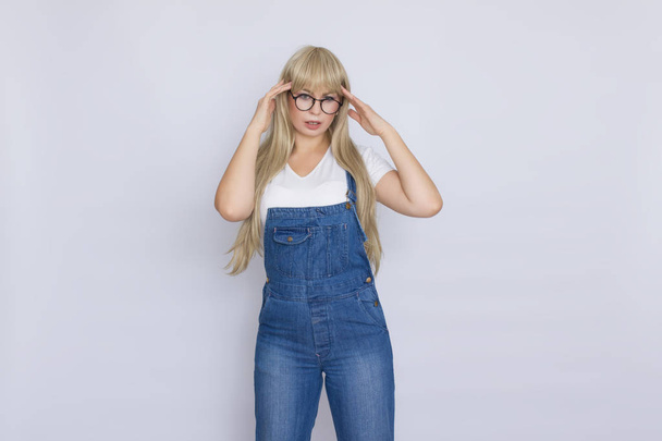 Sweet beautiful blond woman with very long hair in blue jeans overalls and glasses over grey background - Fotografie, Obrázek
