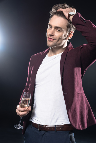 handsome young man in stylish blazer holding champagne glass and looking at camera on black - Foto, Imagem