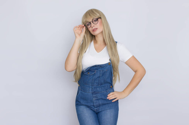 Sweet beautiful blond woman with very long hair in blue jeans overalls and glasses over grey background - Zdjęcie, obraz