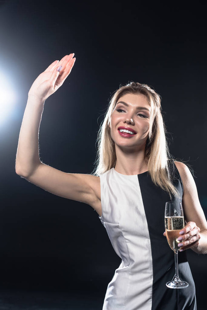 beautiful young woman holding glass of champagne and waving with hand on black - Foto, Imagen