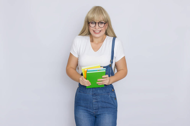 Studio portrait of a beautiful young blonde woman in blue denim overalls and glasses holding books in her hands over grey background - Photo, Image