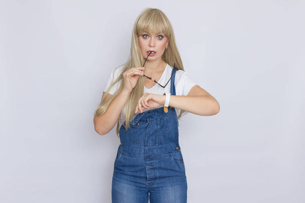 Cute young woman blonde with long hair in a denim jumpsuit and glasses. The girl has a white watch on her hand. She posing over grey background - Valokuva, kuva