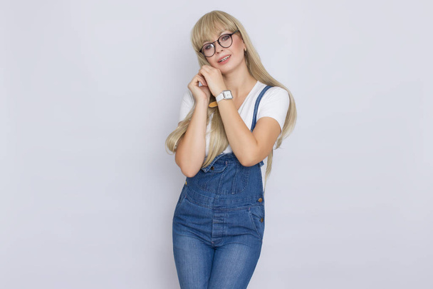 Cute young woman blonde with long hair in a denim jumpsuit and glasses. The girl has a white watch on her hand. She posing over grey background - Φωτογραφία, εικόνα