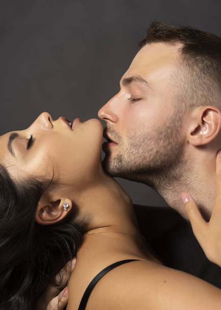 Closeup of a beautiful couple. A man sensually touches his lips to the chin of a girl whose shoulder is bare. Casual fashionable style. Lifestyle, fashion, commercial design. Gray background. - Fotó, kép