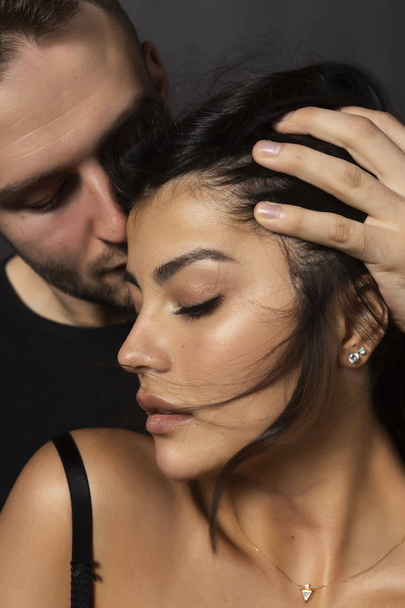 Closeup of a beautiful couple. A man sensually touches his lips to the forehead of a girl whose shoulder is bare. Casual fashionable style. Lifestyle, fashion, commercial design. Gray background. - 写真・画像