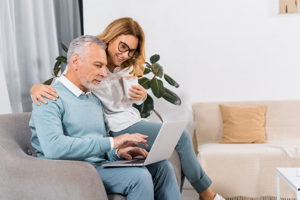 a középső idős ember használ laptop mosolygó felesége kávé otthon ül míg oldalnézete - Fotó, kép