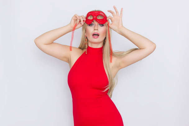 Portrait of a beautiful young blonde woman with long hair in a red evening dress. Girl puts on a red carnival mask over white background - Photo, image