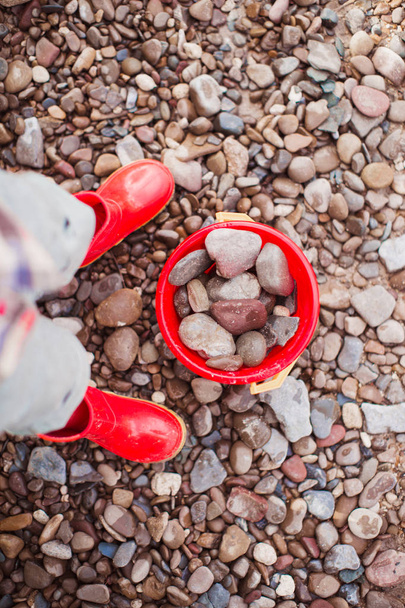 The little boy standing outdoor near the red bucket with the stones - 写真・画像