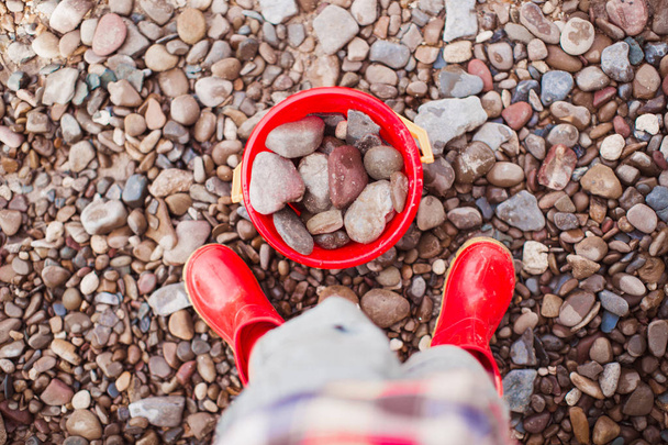 The little boy standing outdoor on the stones near the red bucket with the stones - 写真・画像