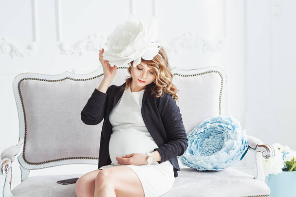 Retrato de uma jovem mulher grávida bonita feliz com flor branca grande nela tinha sentado no sofá branco e segurando seu estômago
 - Foto, Imagem