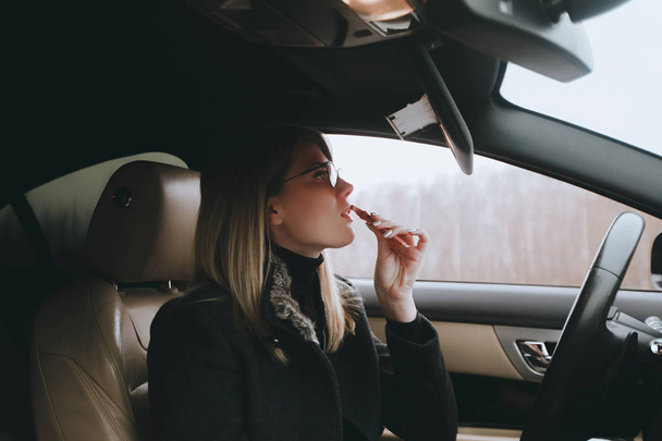 Νεαρή ελκυστική ξανθιά βάφει τα χείλη σε καθρέφτη αυτοκινήτου. Τρόπο ζωής και τα αυτοκίνητα - Φωτογραφία, εικόνα