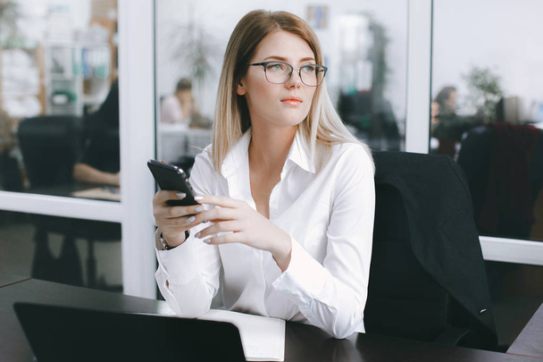 Calm serious young attractive blonde uses phone while working at table in office. Business portrait - Foto, imagen