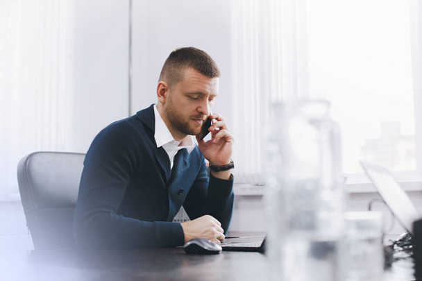 Спокойный серьезный молодой человек пользуется телефоном во время работы за столом в офисе. Бизнес-портрет
 - Фото, изображение