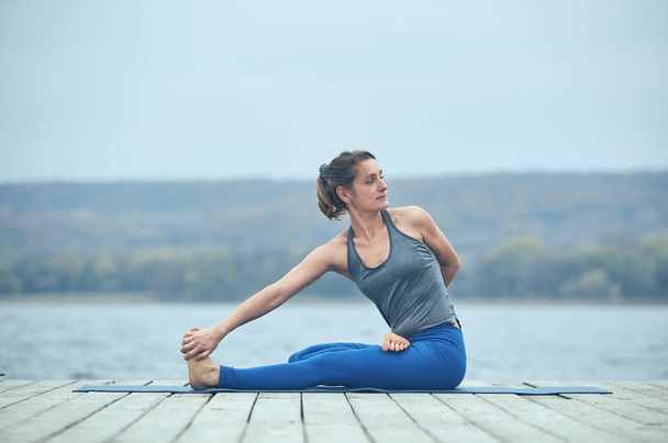 Belle jeune femme pratique le yoga asana Bharadvajasana sur la terrasse en bois près du lac
 - Photo, image