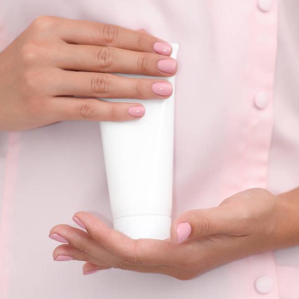 Hand skin care. Female hands holding one white cream tube against body in pink cloth, beautiful beautician woman hands with manicured nails close-up background, shallow DOF, square composition. - Photo, image