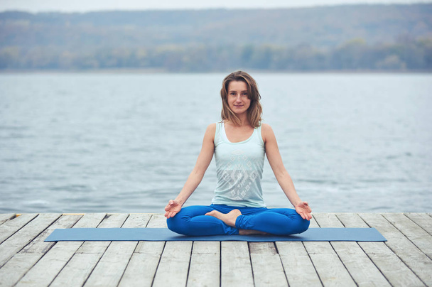 Beautiful young woman practices yoga asana Padmasana - Lotus pose on the wooden deck near the lake - Photo, image