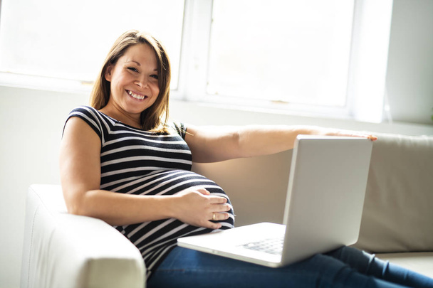 A portrait of a beautiful pregnant woman using laptop lay on the couch - Fotó, kép