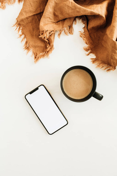 Mug of coffee with milk, brown blanket, smart phone with blank screen mockup on white background. Flat lay, top view. - Photo, image