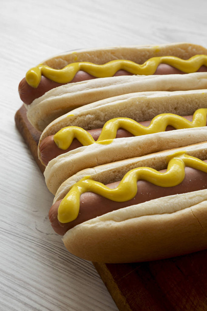 Hot Dog mit gelbem Senf auf Holzbrett auf weißem Holztisch, Seitenansicht. Nahaufnahme. - Foto, Bild