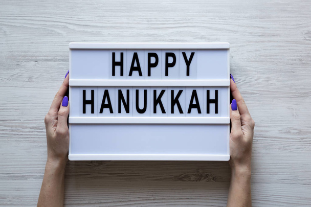 Las manos femeninas sostienen tablero moderno con texto 'Happy Hanukkah' sobre fondo de madera blanca, vista aérea. Desde arriba, plano laico, vista superior
. - Foto, imagen