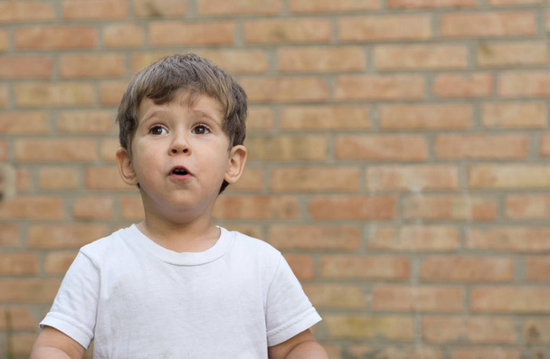 Šťastné dítě chlapeček v bílé tričko vyjadřuje potěšení na prázdné prázdné zdi. Dítě u prázdné cihlová zeď. - Fotografie, Obrázek