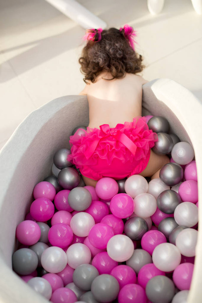 Šťastná holčička sedící v barevných koulích. holčička s barevnými plastovými kuličkami v bazénu, pohled zezadu - Fotografie, Obrázek
