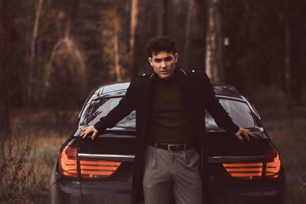 A stylish young man wearing a black coat leaning on a luxury car in the autumn forest - Photo, Image