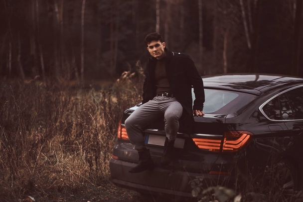 Стильный молодой человек в черном пальто сидит на багажнике автомобиля в осеннем лесу
 - Фото, изображение