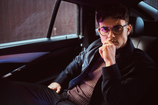 Um jovem pensativo usando óculos sentado no banco de trás do carro
. - Foto, Imagem
