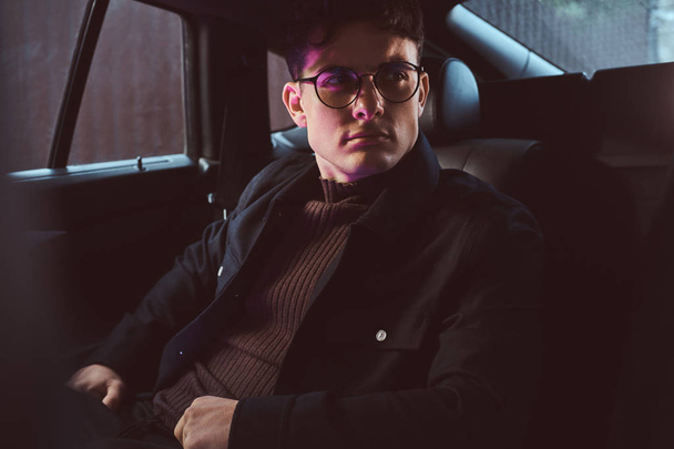 Un joven guapo con gafas sentado en el asiento trasero del coche
. - Foto, imagen