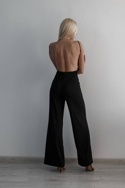 Menina loira bonita elegante em um vestido preto de moda com uma parte traseira aberta está de pé perto da parede branca
 - Foto, Imagem