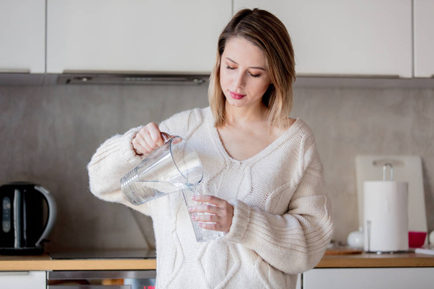 Chica joven en suéter con vaso de agua en la cocina
 - Foto, imagen