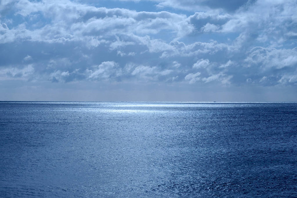 Zee en de lucht in de onderste helft is een kalme diepblauwe zee, aan de horizon is een lijn van wit glinsterende gloeiende licht van de maan in de bovenste helft is de hemel bedekt met pluizige wolken de afbeelding heeft een blauwe gloed uit het maanlicht - Foto, afbeelding