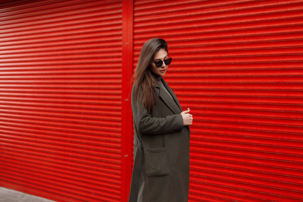 スタイリッシュなサングラスと赤い金属壁の近く、トレンディなファッション グリーン コートに立ってかなり若い魅力的な女性. - 写真・画像