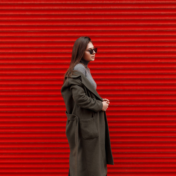 Κομψό όμορφη νεαρή γυναίκα με γυαλιά ηλίου σε ένα μαύρο κομψό παλτό κοντά στον κόκκινο μεταλλικό τοίχο - Φωτογραφία, εικόνα