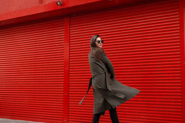 Stylová krásná mladá atraktivní žena s sluneční brýle v moderní zelené kabát v pohybu kovové červené zdi - Fotografie, Obrázek