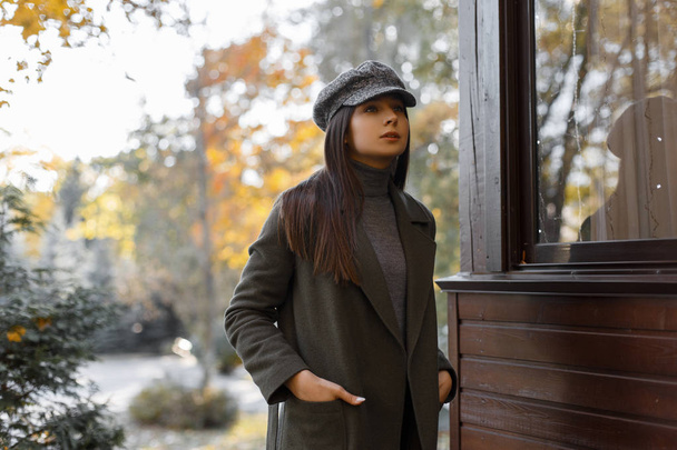 Stylish and young girl in a gray coat and stylish cap stands in the park on an autumn afternoon. Beautiful woman - Φωτογραφία, εικόνα