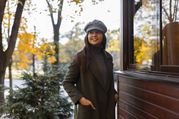 Стильна молода красива жінка з посмішкою у вінтажному капелюсі з зеленим пальто в парку біля дерев'яної будівлі
 - Фото, зображення