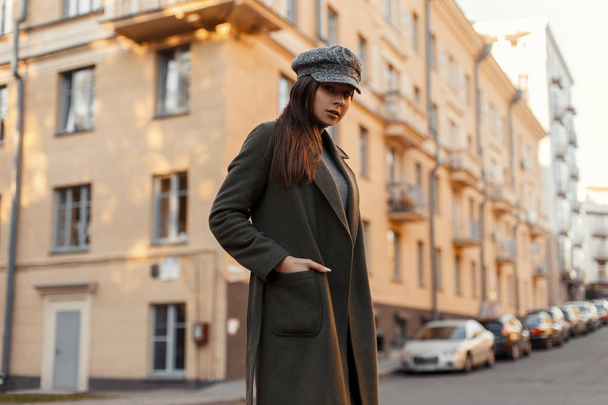 ヴィンテージの帽子とおしゃれなグリーン コートでスタイリッシュな美しい若い魅力的な女性が路上でヨーロッパの都市で歩く  - 写真・画像