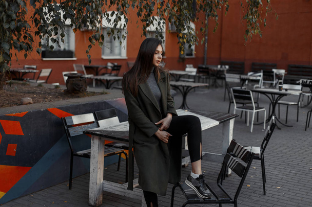 Beautiful and young stylish girl in a vintage coat and black pants is sitting on a table in a modern outdoor cafe against the background of a red wall. Woman hipster. - Valokuva, kuva