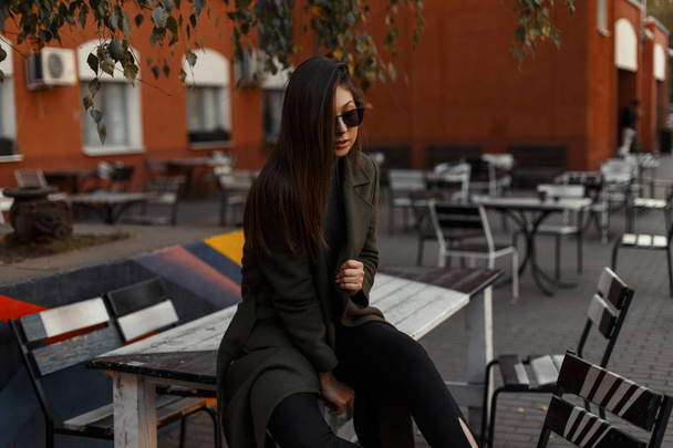 Bella e alla moda ragazza modello bruna in un elegante cappotto verde e occhiali da sole è seduto in un caffè all'aperto. Accessori moda e vestiti alla moda. Tendenze bellezza
. - Foto, immagini