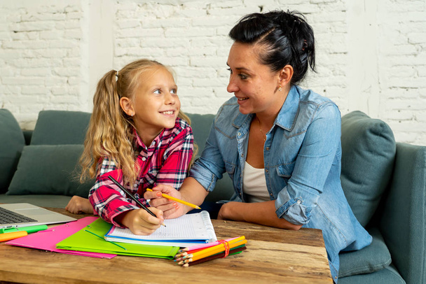 Beautiful mother helping her young blonde cute sweet girl to do homework at home in homework parenting and education concept. - Photo, image
