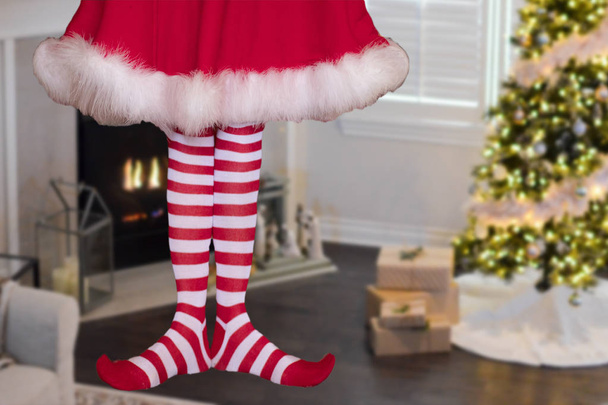 かわいいクリスマスエルフの女の子は、バックグラウンドで贈り物やクリスマスツリーと装飾されたクリスマスリビングルームに立って縞模様のストッキングを身に着けています - 写真・画像