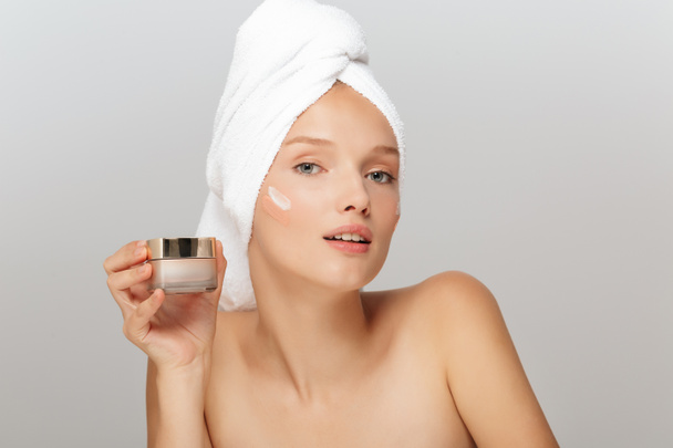 Portrait of young attractive lady with white towel on head without makeup holding face cream in hand thoughtfully looking in camera with cream on cheek over gray background - Photo, Image