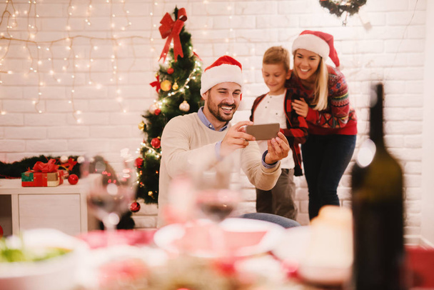 young happy parents and their adorable little son celebrating Christmas at home and taking selfie  - Photo, Image