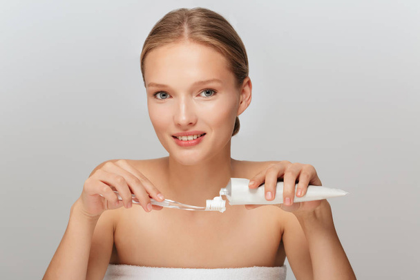 Porträt einer jungen schönen Frau ohne Make-up, die etwas Zahnpasta auf die Zahnbürste legt und verträumt vor grauem Hintergrund in die Kamera schaut - Foto, Bild