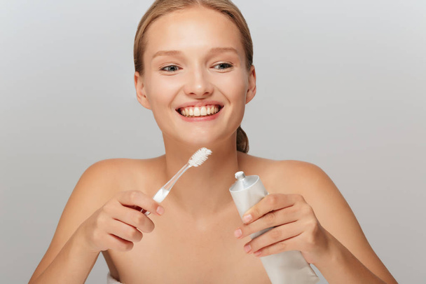 Portrait of young cheerful lady without makeup holding toothpaste and toothbrush in hands joyfully looking aside over gray background - Photo, Image